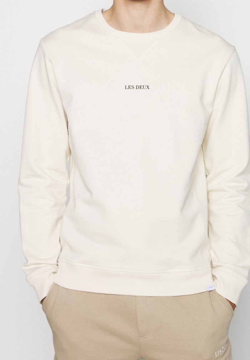 Homme portant un sweat-shirt crème à manches longues avec le texte "LES DEUX" centré sur la poitrine, associé à un pantalon beige, les mains dans les poches.