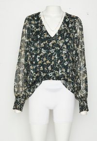 Blouse longue transparente à manches longues avec imprimé floral, poignets smockés et col en V, associée à un short blanc ajusté sur un mannequin.