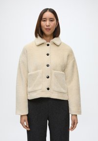 Kreminė dirbtinio kailio striukė su juodais mygtukais, plokščiu apykakle, dviem priekinėm kišenėm ir trumpu dizainu. Minkšta tekstūra ir laisvas siluetas.