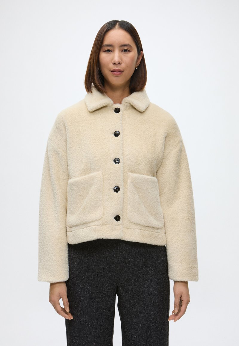 Kreminė dirbtinio kailio striukė su juodais mygtukais, plokščiu apykakle, dviem priekinėm kišenėm ir trumpu dizainu. Minkšta tekstūra ir laisvas siluetas.