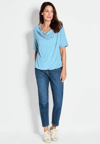 Lichtblauw gedrapeerd topje met een ronde hals, gecombineerd met medium-wash jeans en witte sneakers. De stof lijkt zacht en glad te zijn.