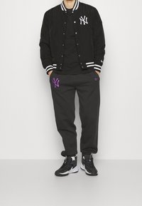 Black varsity jacket with white stripes and NY logo, paired with black sweatpants featuring purple NY logo. Black sneakers.