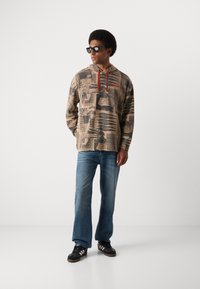 Long-sleeve beige hoodie with graphic prints in black and red, featuring a drawstring hood; paired with light blue jeans and black sneakers.