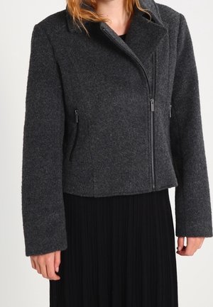 Femme portant une veste en laine gris foncé avec fermeture éclair, col et manches longues, sur une jupe plissée noire.
