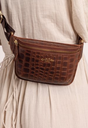 Braune strukturierte Leder-Gürteltasche mit goldenem Reißverschluss und Logo, getragen über beigem Leinenkleid mit gebundener Taille.