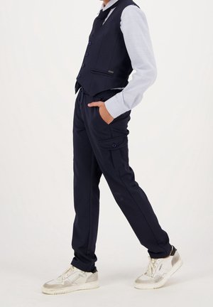 Person wearing navy blue vest and cargo pants with white long-sleeve shirt, hands in pockets, and white sneakers, standing sideways.