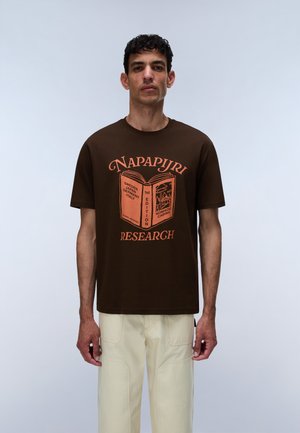 Homme portant un t-shirt Napapijri marron avec un texte graphique orange et un pantalon beige, debout devant un fond clair uni.
