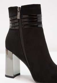 Bottine en daim noir avec fermeture éclair latérale, trois brides noires brillantes près du haut et un talon bloc argenté réfléchissant.