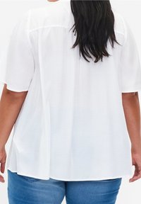 Femme aux longs cheveux bruns portant un chemisier blanc à manches courtes amples et un jean bleu, debout dos à la caméra.