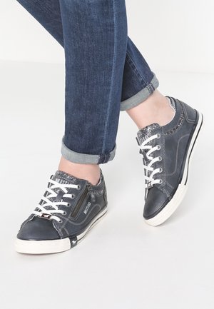 Osoba w ciemnoniebieskich dżinsach i ciemnoszarych niskich trampkach z białymi sznurowadłami i bocznym zamkiem, stojąca na jasnej podłodze.