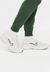 Λευκά αθλητικά παπούτσια Nike με λεπτομέρειες από διχτυωτό και δερμάτινο υλικό, με σκούρο πράσινο swoosh. Συνδυασμένα με λευκές κάλτσες και πράσινα φόρμα.