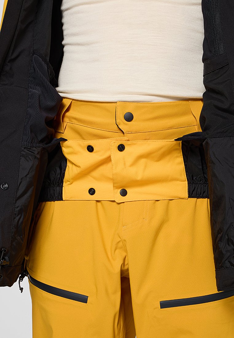Pantaloni da sci impermeabili gialli con vita aderente, chiusura a snap e dettagli neri. Il tessuto è testurizzato e resistente, progettato per il freddo.