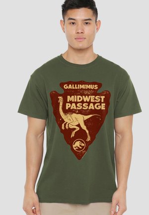 Junger Mann trägt ein olivgrünes T-Shirt mit brauner Dinosaurier-Grafik und Text "Gallimimus Midwest Passage."