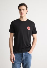 Black cotton t-shirt featuring a small round red logo with "HUGO" printed inside. Short sleeves and a crew neckline. Worn with blue jeans.
