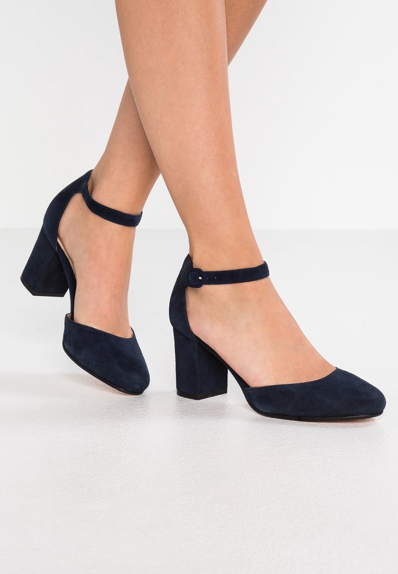 Jambes portant des talons blocs en daim bleu marine avec des brides à la cheville, debout sur un sol clair contre un fond blanc uni.