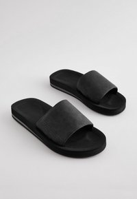 Zwarte slides met een textuur op het bovenste riempje met perforaties, een effen zwarte buitenzool en een licht verhoogd zoolontwerp.