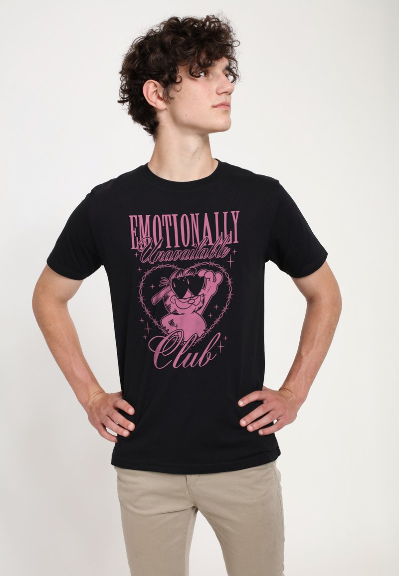 Jeune homme aux cheveux bouclés portant un t-shirt noir avec l'inscription « Emotionally Unavailable Club » avec un personnage de dessin animé et des lunettes en forme de cœur.