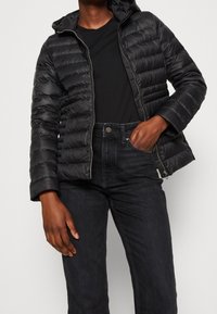 Doudoune noire avec une fermeture éclair à l'avant, une capuche et des poignets côtelés. Portée par-dessus une chemise noire et assortie avec un jean foncé.