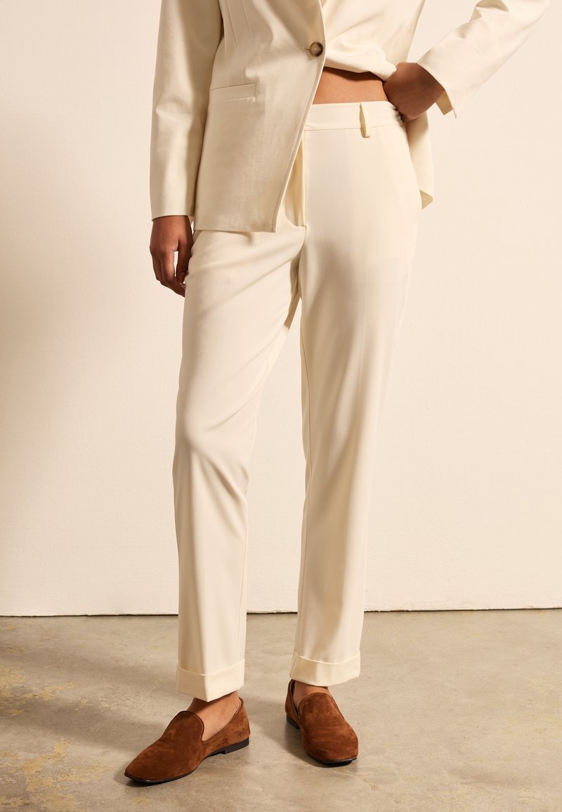 Cremefarbene, maßgeschneiderte Hose mit glatter Textur, flachem Schnitt, seitlichen Taschen und umschlagenen Bündchen, kombiniert mit braunen Slippern.
