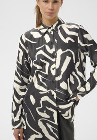 Naine, kes kannab pika varrukaga musta nööbitavat särki suurte abstraktsete valgete mustritega, seisab ühe käega puusal.