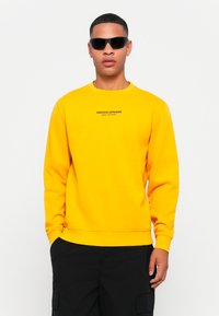 Bright yellow sweatshirt with round neckline, ribbed cuffs, and black text logo on the chest. Model wears black sunglasses.