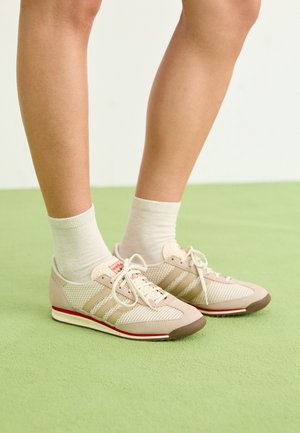 Béžové sportovní boty vyrobené z jemné síťoviny a semiše, design se třemi pruhy, lehká gumová podešev a červené akcenty, nošeny s bílými ponožkami.