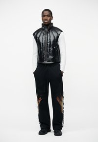 Mężczyzna w czarnej, błyszczącej, bezrękawnikowej kamizelce na zamek, noszący pod nią biały, długi rękaw oraz czarne spodnie z wzorem płomieni i napisem po bokach, stojący z rękami w kieszeniach.