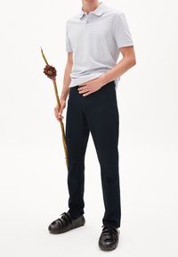 Ljusgrå pikétröja med korta ärmar och en liten logotyp, kombinerad med mörkblå byxor och svarta snörskor. Modellen håller i en växtstjälk.