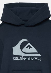 Marineblauer Kapuzenpullover mit einem großen grauen Quiksilver-Logo, das ein Bergmotiv innerhalb einer Welle zeigt, und einer Kängurutasche. Weicher Stoff.