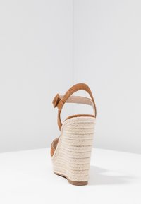 Sandale compensée en daim couleur camel avec plateforme en jute tressée et bride à la cheville, vue de dos sur une surface blanche avec un fond gris uni.