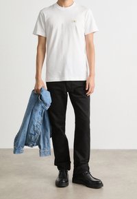 Persona che indossa una maglietta bianca a maniche corte, pantaloni neri, scarpe nere, tiene in mano una giacca di denim blu con la mano sinistra, in piedi su un pavimento grigio contro un muro bianco.