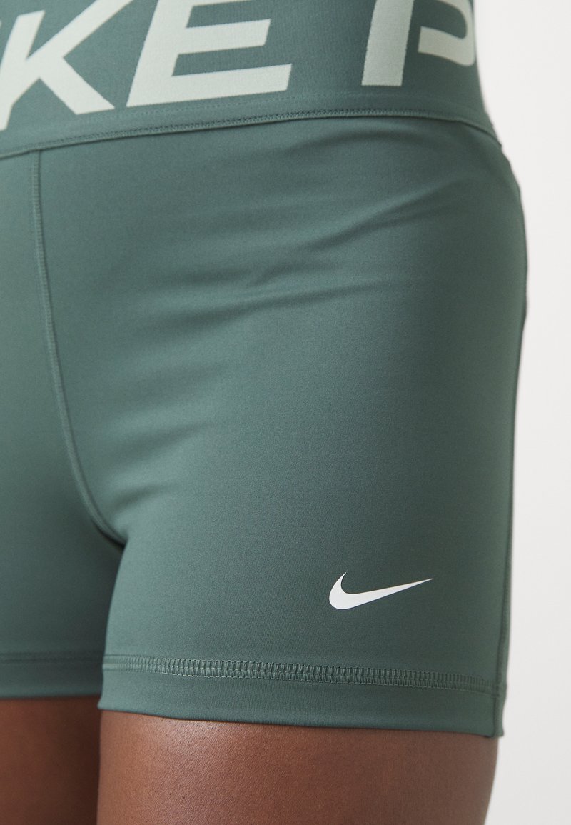 Shorts Nike en tissu vert foncé. Dotés d'une ceinture logo grise et d'un petit swoosh blanc sur la jambe. Texture lisse, design ajusté.