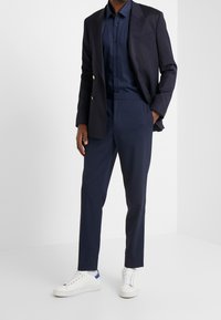 Marineblazer met gouden knopen, gecombineerd met een marineblauw overhemd en op maat gemaakte marineblauwe broek. Witte sneakers met blauwe accenten maken de outfit compleet.