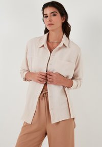 Lichtbeige button-up shirt van zachte, textuurrijke stof. Heeft een kraag, lange mouwen en een borstzak. Gepaard met tan kleurige broeken.