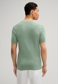 Maglione a maniche corte di colore verde chiaro, in maglia tessuta. Presenta un colletto rotondo e un orlo dritto. Indossato con pantaloni di colore chiaro.