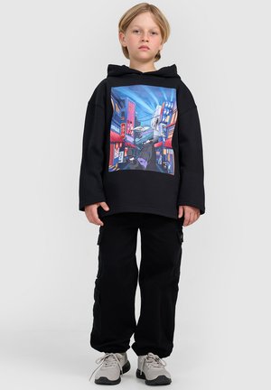 Dziecko z krótkimi blond włosami ubrane w czarną bluzę z kapturem z kolorową miejską grafiką, czarne spodnie cargo i szare sneakersy.