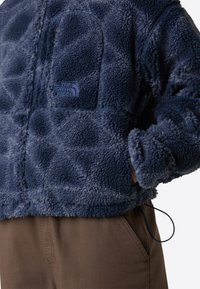Blå fleecejakke med en teksturert, fluffy overflate, glidelåslomme på brystet og logo-akcent. Båret over brune bukser med snor.