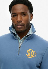 Pulóver azul de forro polar con media cremallera, que presenta un logo dorado, y una camiseta con cuello y rayas debajo. Textura suave y corte relajado.