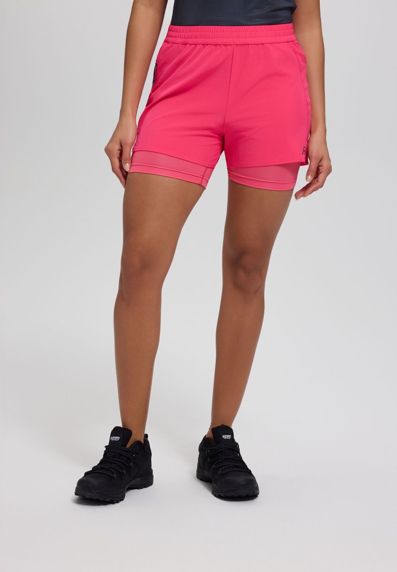 Kvinde iført lyserøde lagdelte træningsshorts og sorte vandresko, stående mod en ensartet lys baggrund.