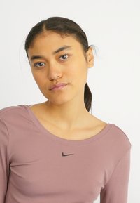 Hosszú ujjú, mályva színű sportpóló bordázott textúrával és fekete Nike logóval a mellrészen. Széles nyakkivágással és testhez simuló ujjal rendelkezik.