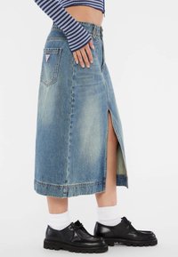 Lichte blauwe denim midi-rok met een voorknos, voorzien van een klassiek vijf-pocketdesign en contrasterende stiksels. G gedragen met zwarte schoenen en sokken.