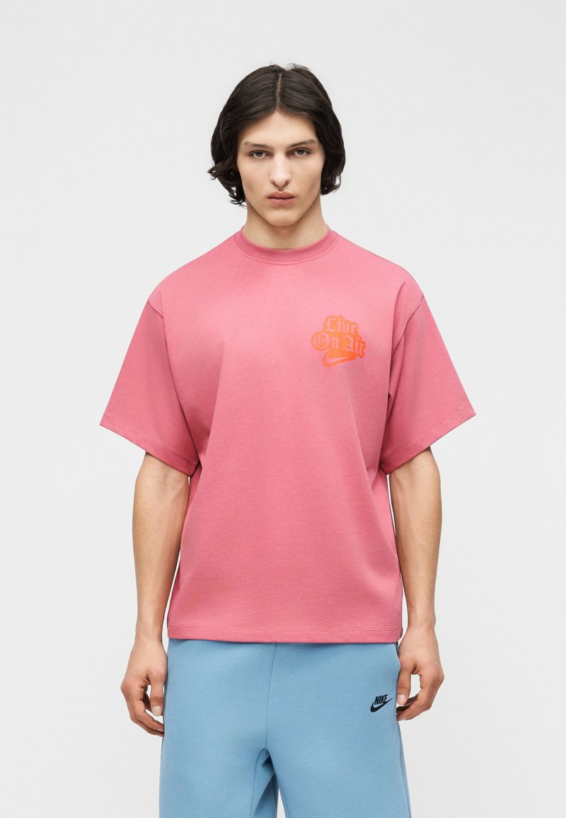 Młody mężczyzna o krótkich czarnych włosach, ubrany w różowy t-shirt Nike z napisem "Live On Air" oraz jasnoniebieskie spodnie dresowe Nike, stoi na tle jednolitego tła.