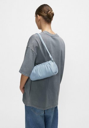 Kobieta z włosami upiętymi w kok, ubrana w luźny szary t-shirt i niebieskie dżinsy, nosi jasnoniebieską marszczoną torbę na ramię na prawym ramieniu.