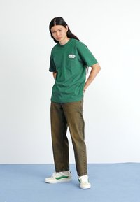 Green short-sleeve t-shirt with a white logo, paired with brown pants and white sneakers featuring green accents, standing on a blue surface.