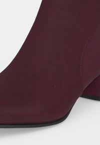 Gros plan d'une bottine en suède rouge foncé avec un talon bloc et une semelle noire sur un fond clair.