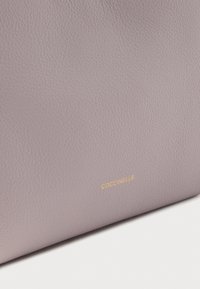 Pehmeä laventelinvärinen teksturoidun nahkapinta, jossa pieni kullanvärinen "COCCINELLE"-logo kohopainettuna lähellä alareunaa.