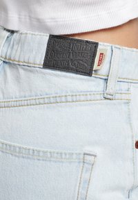 Jeans de mezclilla azul claro con una etiqueta de cuero negro en la cintura, que presentan un detalle de costura amarilla y un bolsillo trasero visible.