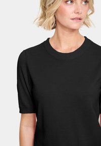 Haut noir à manches courtes avec un col rond. Le tissu semble lisse et côtelé, avec une coupe ajustée et sans motifs ni accents visibles.