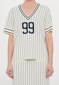 Bílo-námořnický pruhovaný krátký rukáv pletený top s výstřihem do V, s číslem "99" v režném námořnickém písmu a vroubkovanými manžetami.