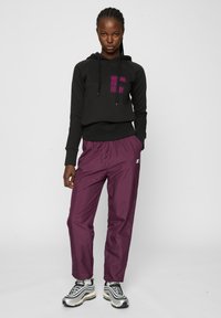 Mujer de pie mirando hacia adelante, vistiendo una sudadera negra con texto rosa y pantalones deportivos morados con zapatillas blancas y negras.